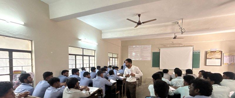 A trainer standing among rows of students seated at desks in a classroom, with a projection screen on the wall behind.
