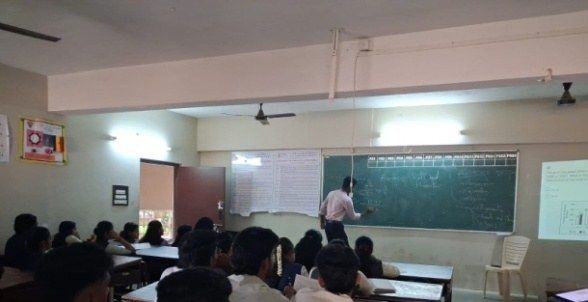 A man writes on a green chalkboard at the front of a classroom while students sit at desks facing him.