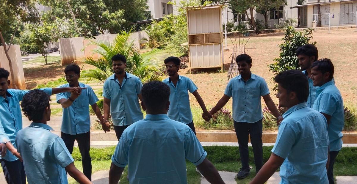 Students in pale blue shirts stand in a ring, holding hands, on a garden lawn with trees and a small hut behind them.