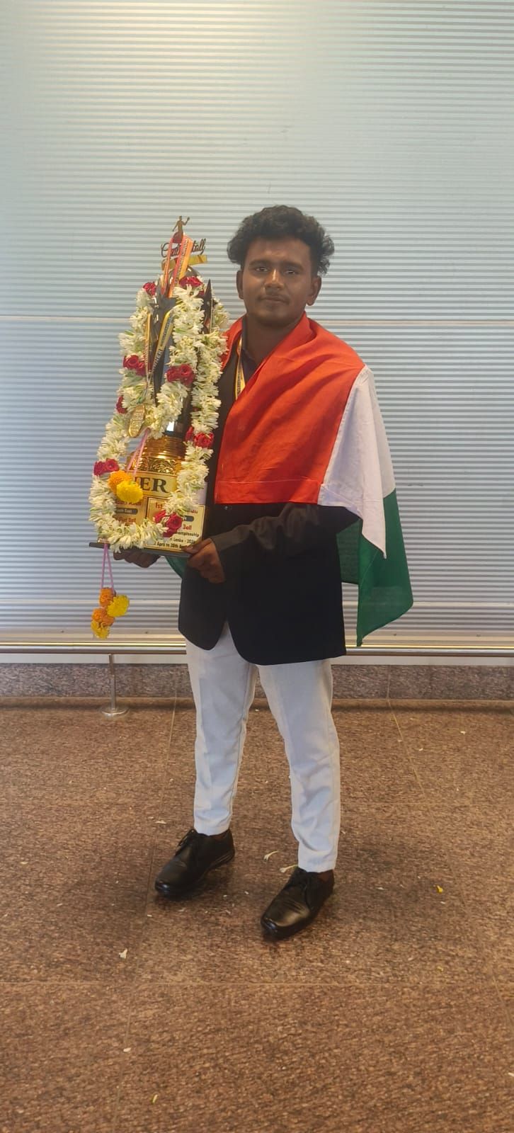 A young man draped in the Indian flag holding a garlanded trophy in an airport hall