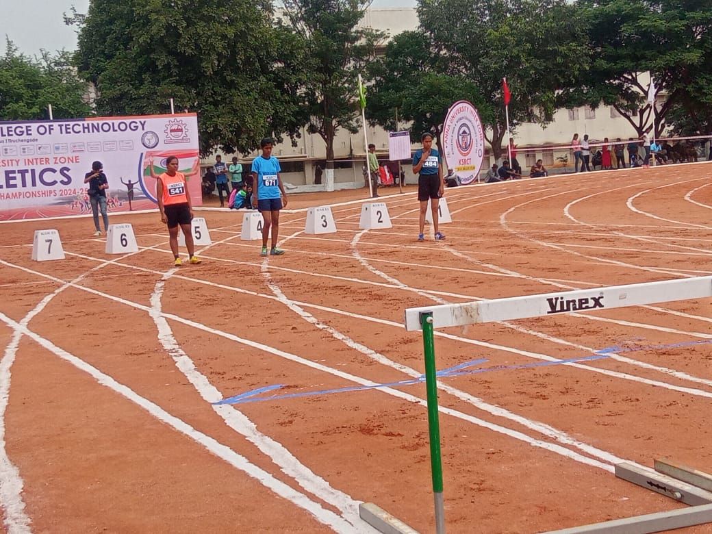 Athletes at the start of a track race on a red track, a hurdle in the foreground and a banner for an inter-zone athletics championship behind them