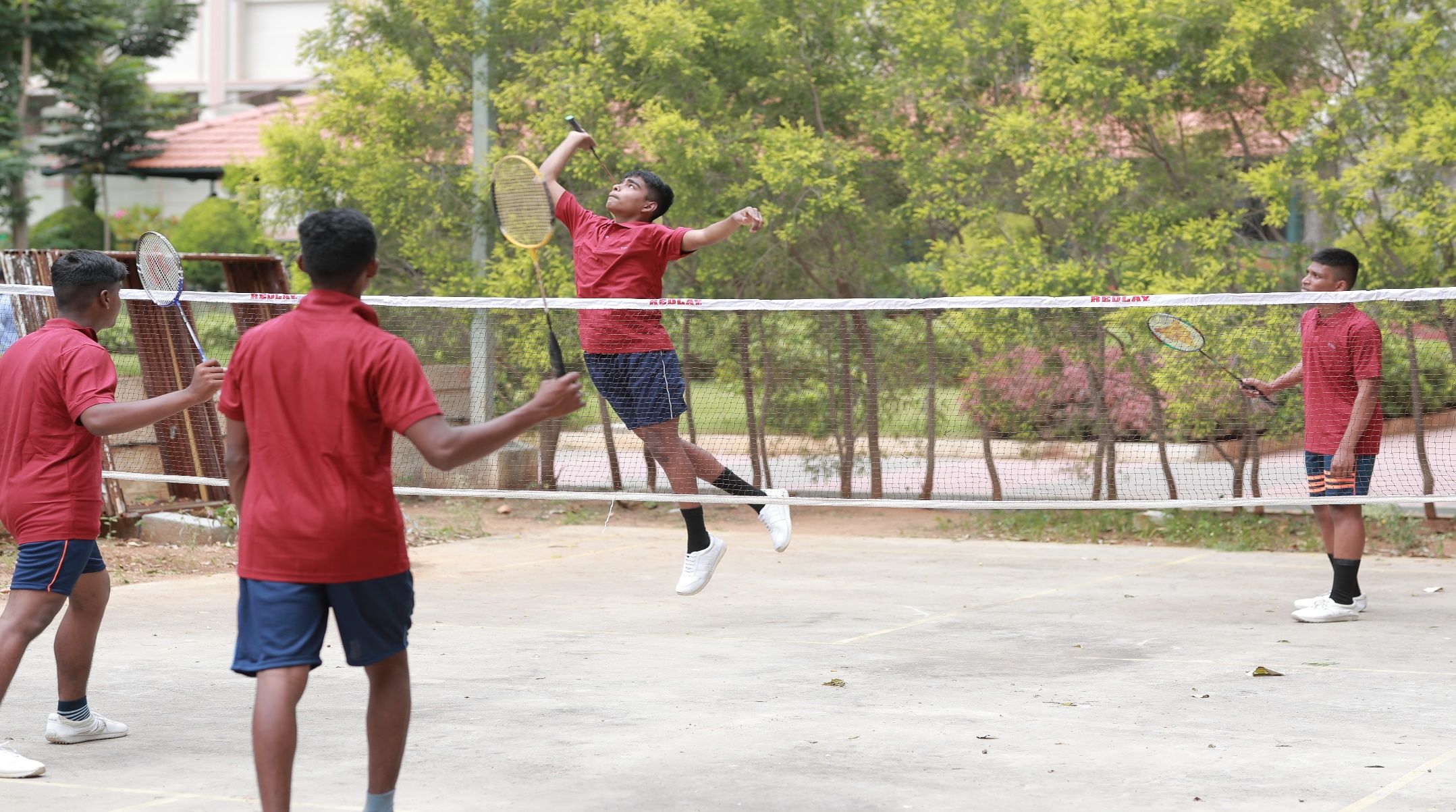 A player in a red shirt leaping to smash at the net of an outdoor badminton court, other players in red and blue shirts around him