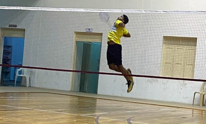 A badminton player in a yellow shirt leaping for a smash at the net of an indoor court