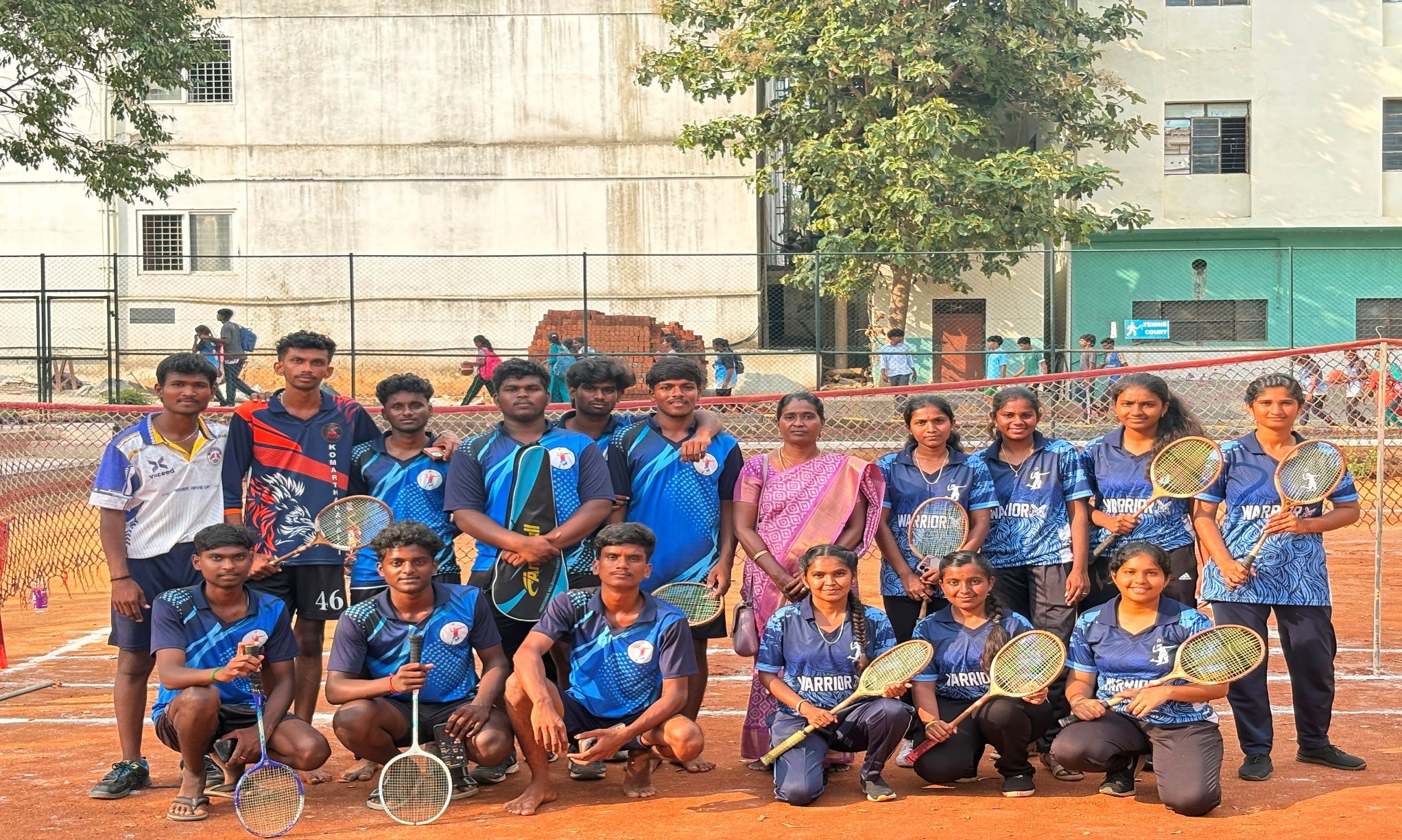 A team of young men and women in blue jerseys posing with badminton rackets on a clay court, a woman in a pink sari among them, a net and trees behind