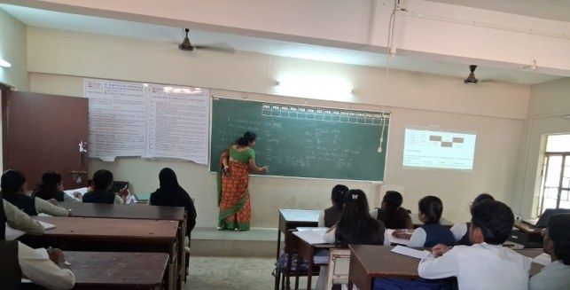A woman in an orange sari writes on a green board at the front of a classroom while students sit at desks and a projected slide shows at the right.