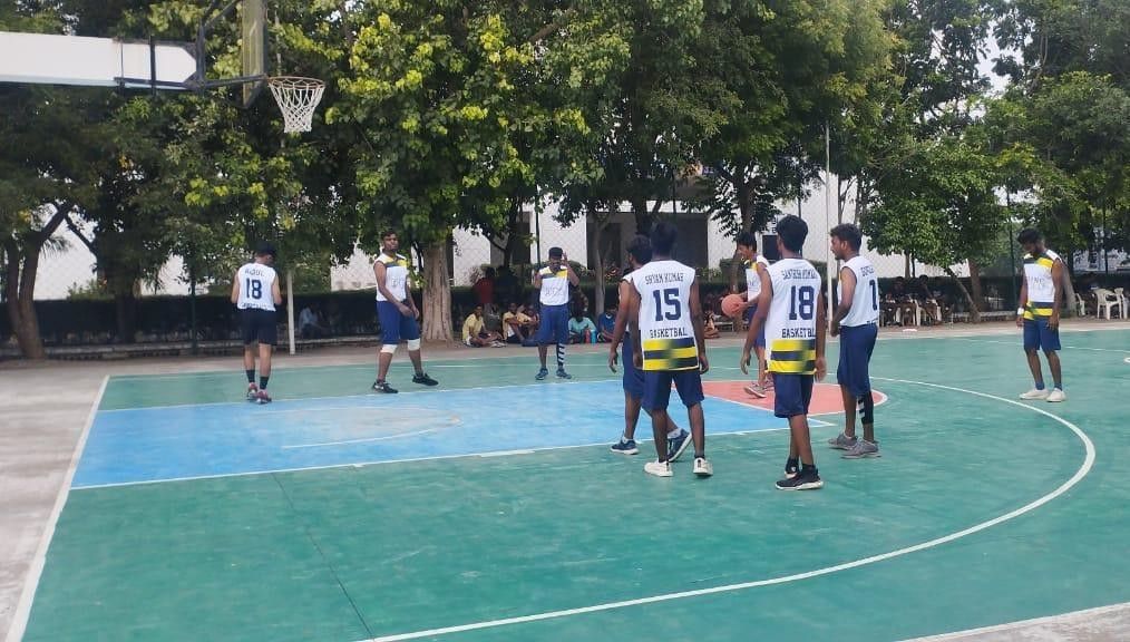 Players in white and navy jerseys contesting a basketball game on a green outdoor court under trees