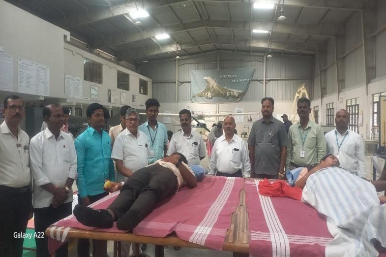 A donor lying on a camp bed giving blood in a large hall, staff and volunteers standing behind