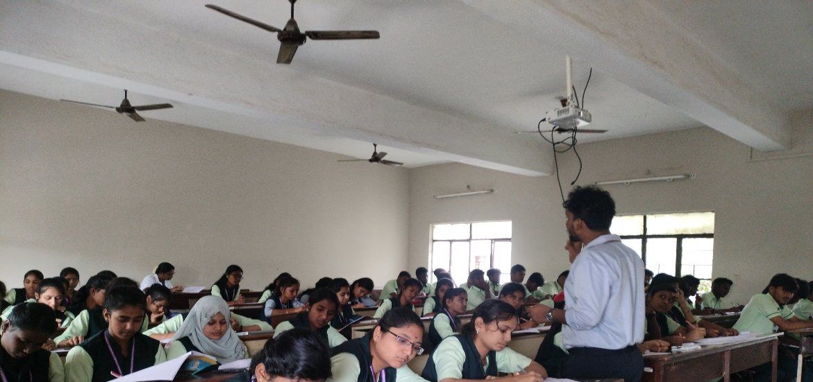 A man in a white shirt stands among students seated at crowded desks in a classroom, with a ceiling projector and a window behind them.