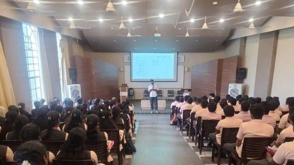 A man stands at the front of a large lecture hall beneath a projection screen while rows of students sit facing him.
