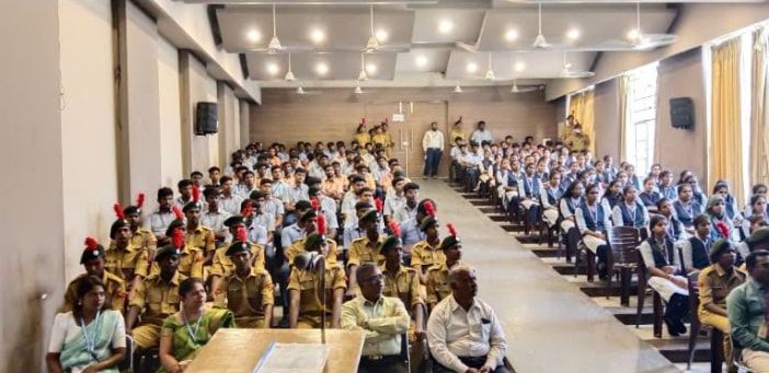 Rows of cadets in khaki uniform and red-plumed berets seated in a hall, with a crowd of students seated beyond them