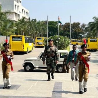 Two cadets in uniform walking with an officer and another man across the college forecourt, yellow buses behind them