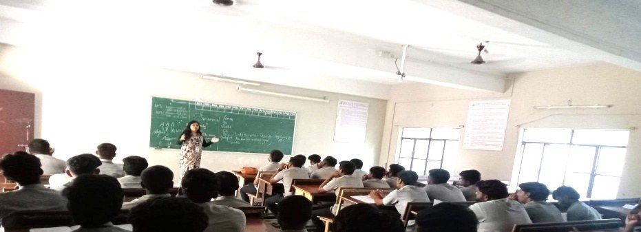 A speaker at a green chalkboard in a large classroom full of seated students.