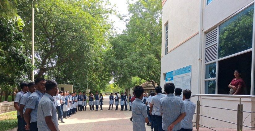 Students stand in a wide circle on a paved path between trees, beside a college building.