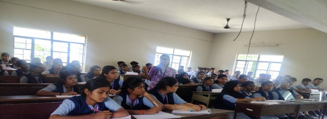 Students in blue-and-white uniform sit writing at long desks in a classroom, with windows and ceiling fans behind them.