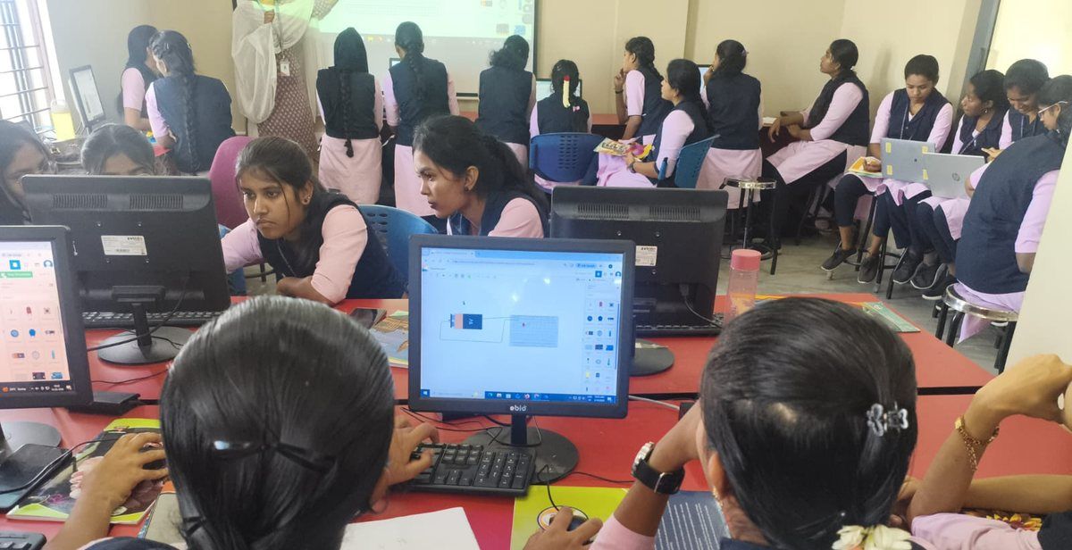 Students in pink and navy uniforms work at computer workstations on red tables, a circuit diagram on the screen in the foreground, while others sit in a group at the back.