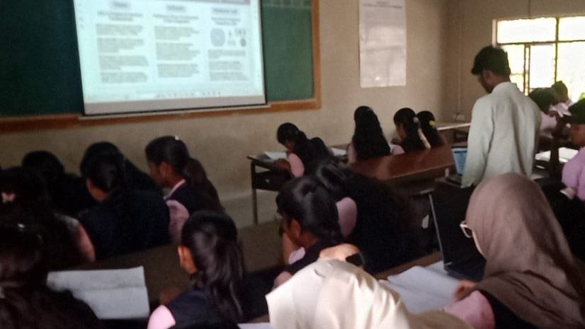 Students in navy and pink uniform sit writing at desks in a classroom while a man in a white shirt stands near a projection screen at the top right.