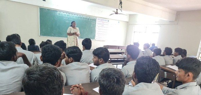 A speaker standing at the front of a classroom beside a green chalkboard while students seated at desks listen.