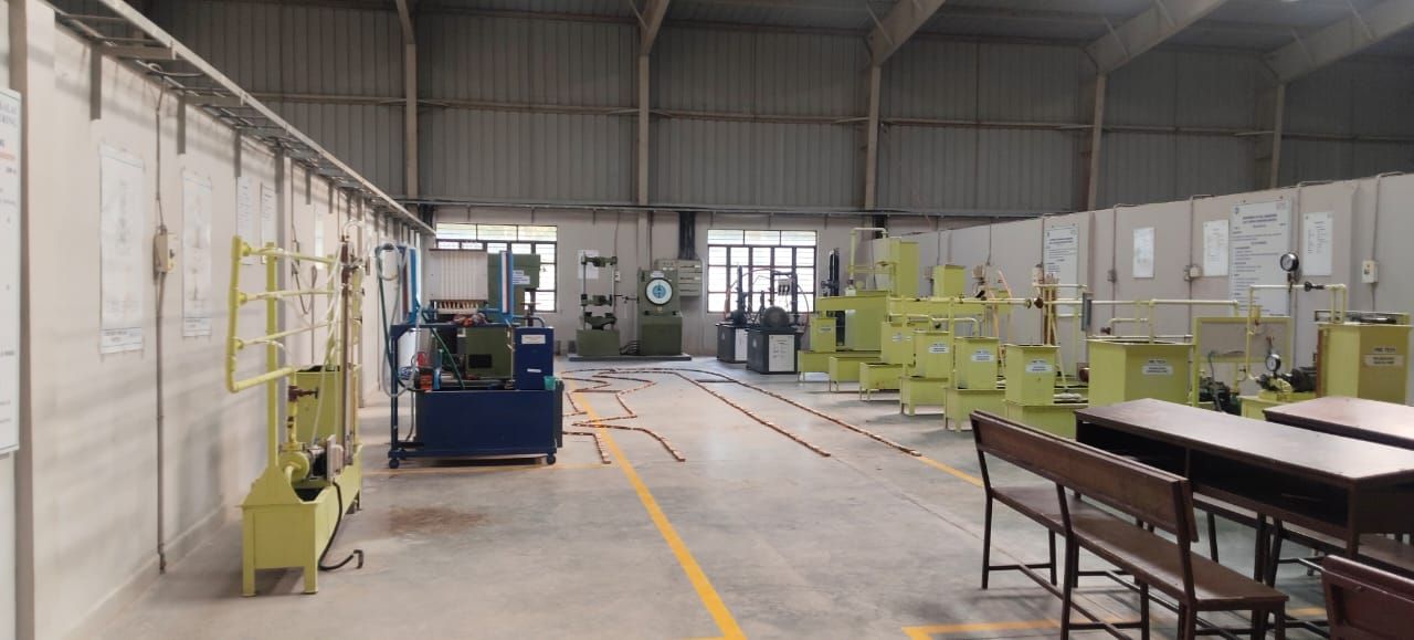 A tall steel-framed hall with yellow-green rigs and pipework along the right-hand wall, a blue machine on the left, more machines at the far end and wooden benches in the right foreground.