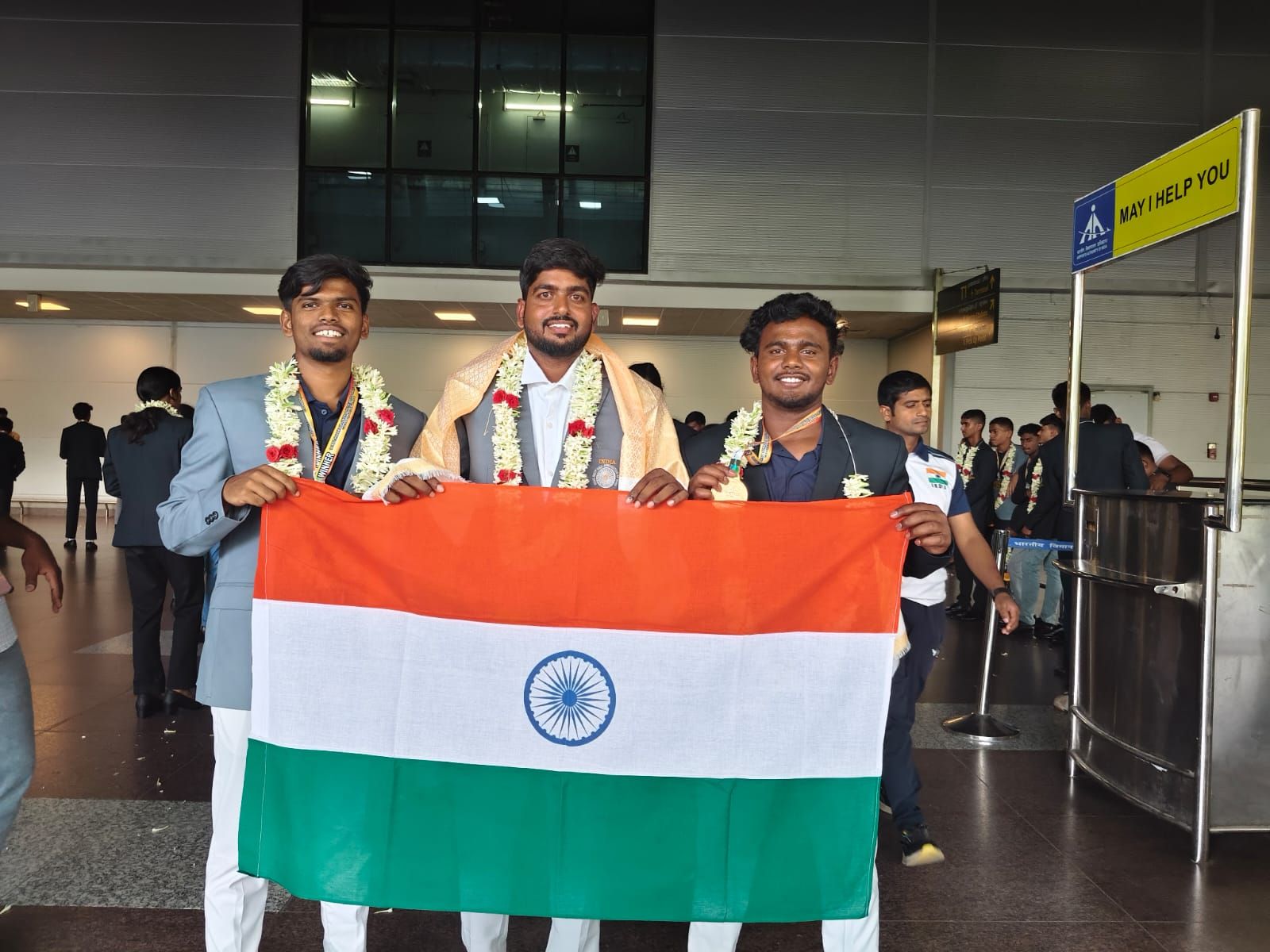 Three young men in flower garlands, medals round their necks, holding the Indian flag between them in an airport hall, with more garlanded people behind them