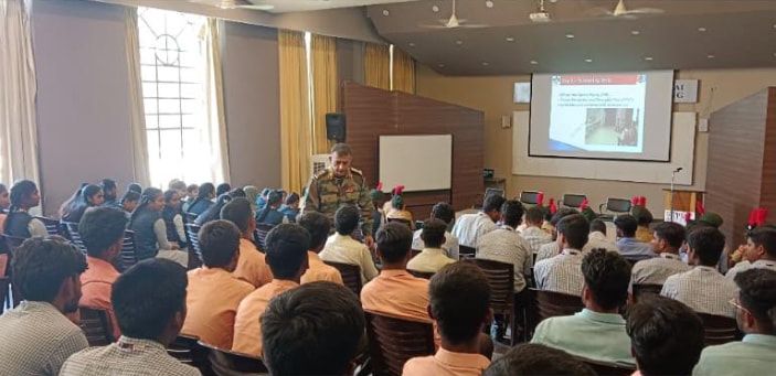 An officer addressing a hall full of students and cadets, a presentation on the screen behind