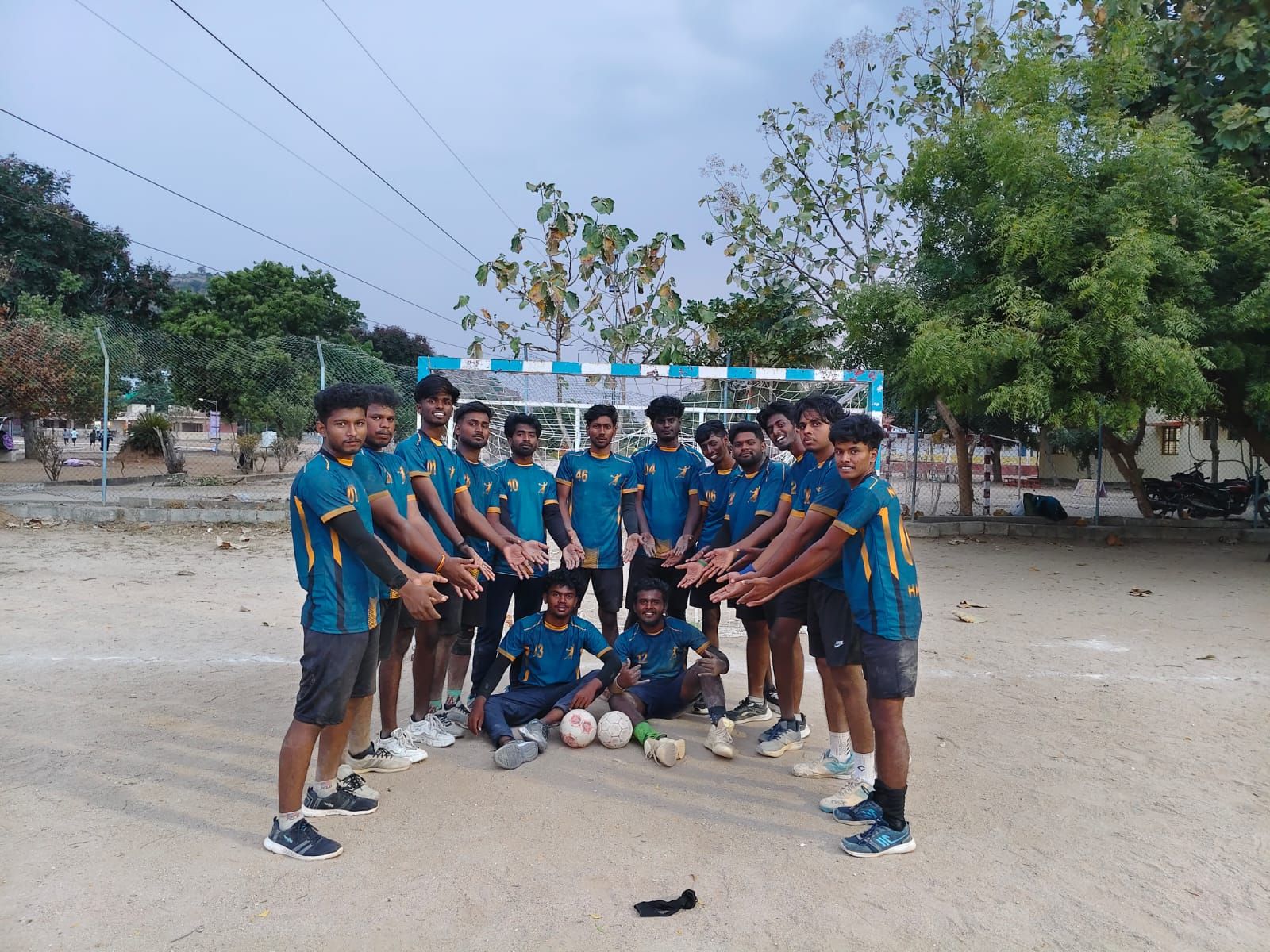 A team in teal jerseys standing in a ring with their hands together over a handball and two more balls on a sandy ground, a goal and trees behind them