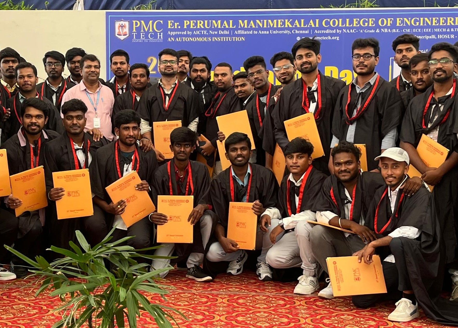 PMC Tech graduates in black gowns and red sashes holding orange folders, posed in front of the college banner