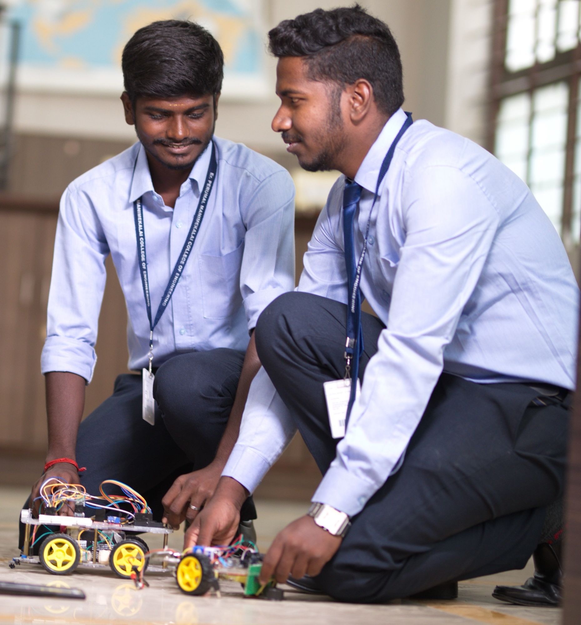 Two PMC Tech students kneeling on the floor, building a small robot car with motors and wiring