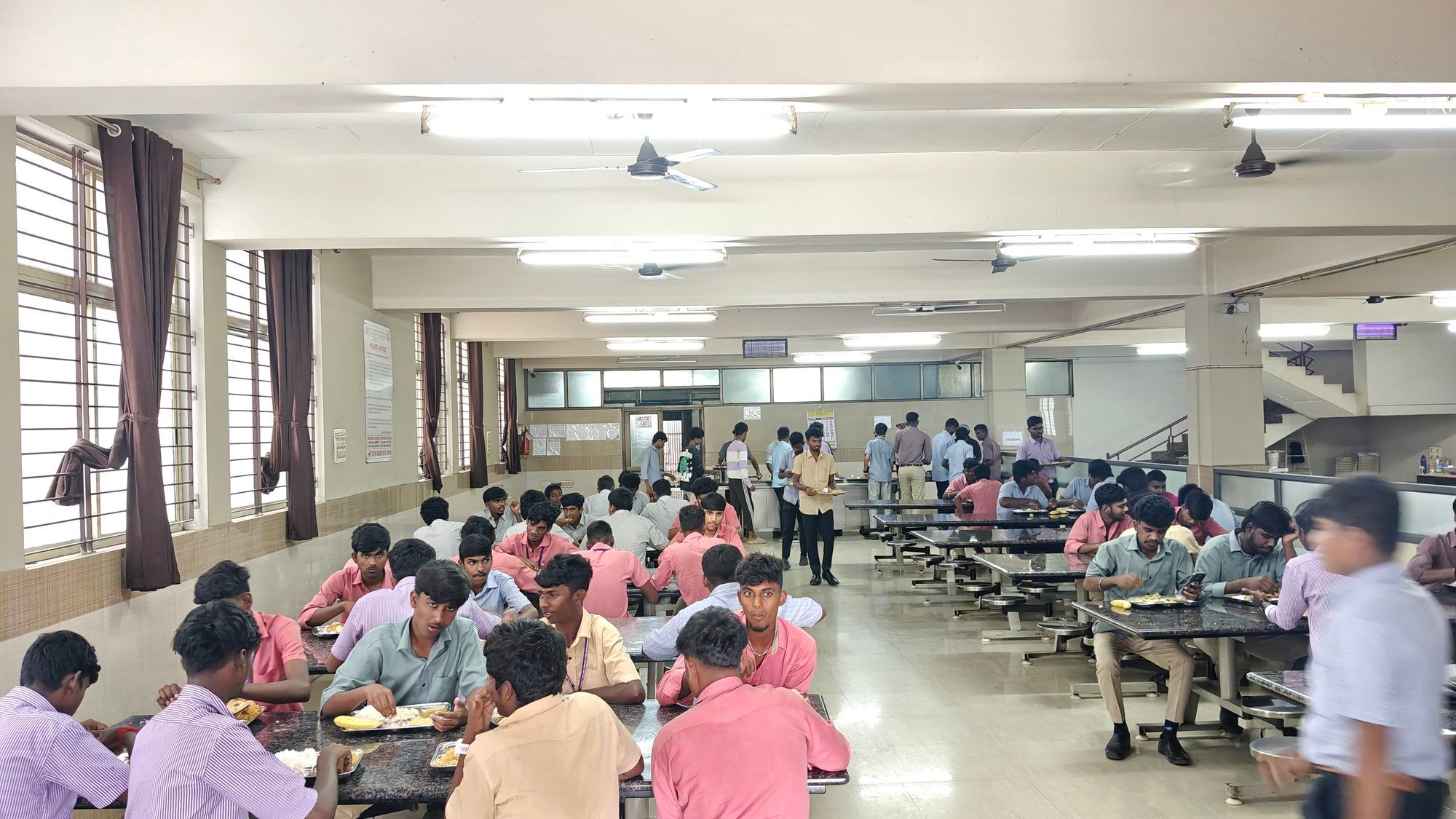 Students eating at long tables in a large hostel dining hall, curtains at its windows