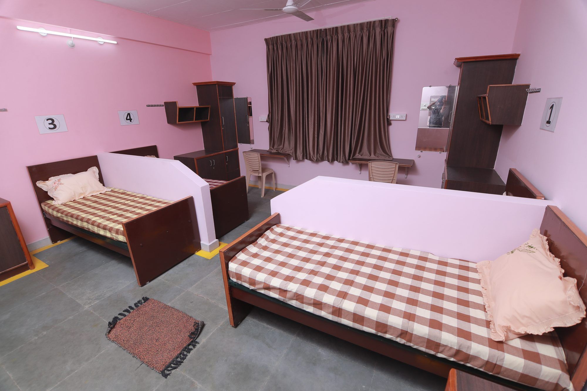 A hostel room with two single beds in checked bedspreads, wooden cupboards and a curtained window