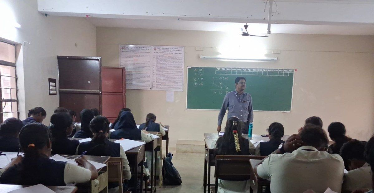 A man in a blue shirt stands at the front of a classroom beside a green board while students sit writing at desks in the foreground.