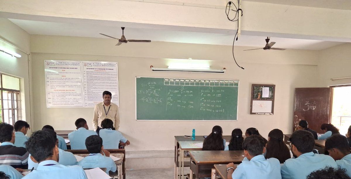 A man stands at a green chalkboard at the front of a classroom while students in pale blue shirts sit at desks facing him.