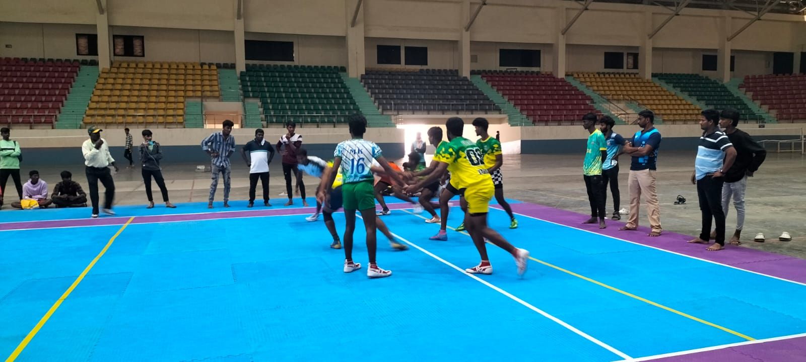 Players from two sides locked together in a kabaddi match on a blue mat in an indoor stadium, onlookers standing round the mat