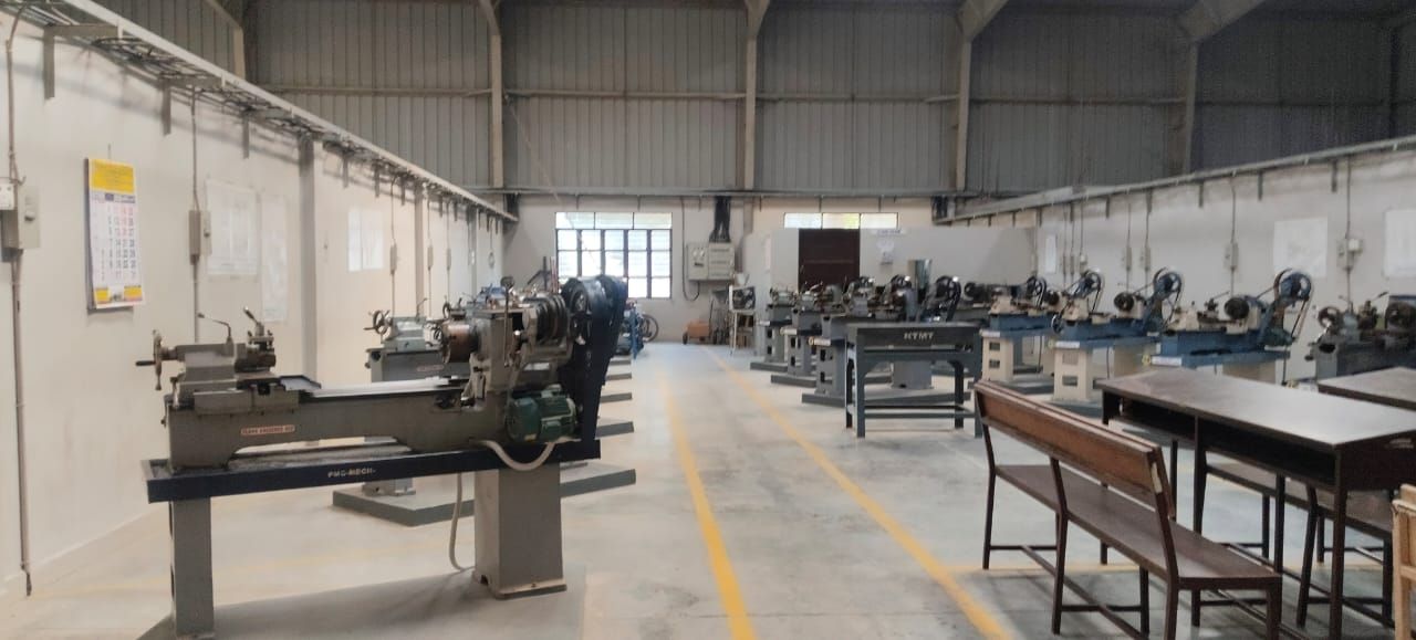 A tall workshop hall with lathes lined up on plinths on both sides of a yellow floor line, and wooden benches in the right foreground.
