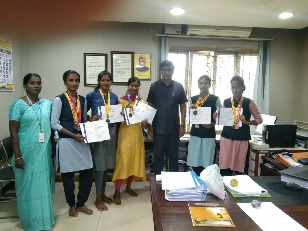 Five students holding certificates, medals round their necks, with a woman and a man beside them in an office