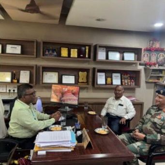 An officer in uniform seated at the end of a table in an office with two other men, shelves of awards behind them