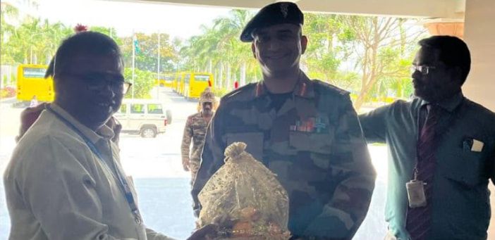 An officer in uniform receiving a wrapped gift from two men at a building entrance, palm trees and yellow buses behind them