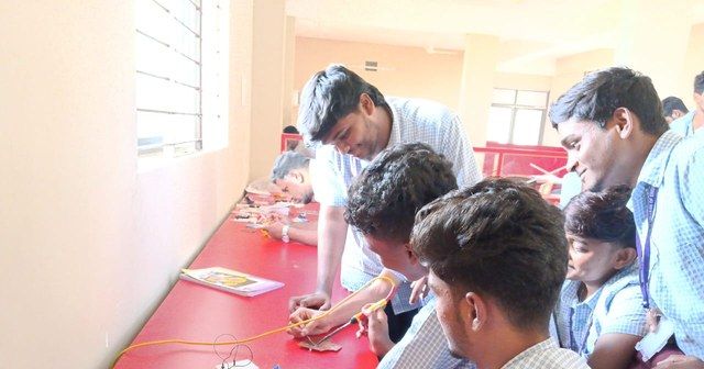 Students bending over a red workbench to work on a circuit of wires while an instructor guides them.