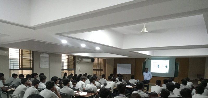 Rows of students seated in a large lecture hall facing a presenter beside a projection screen.