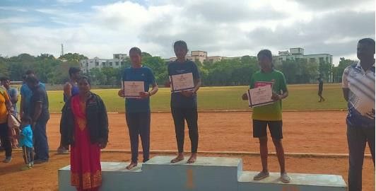 Three athletes on a podium holding certificates, with a woman in a sari and a crowd beside them on a running track