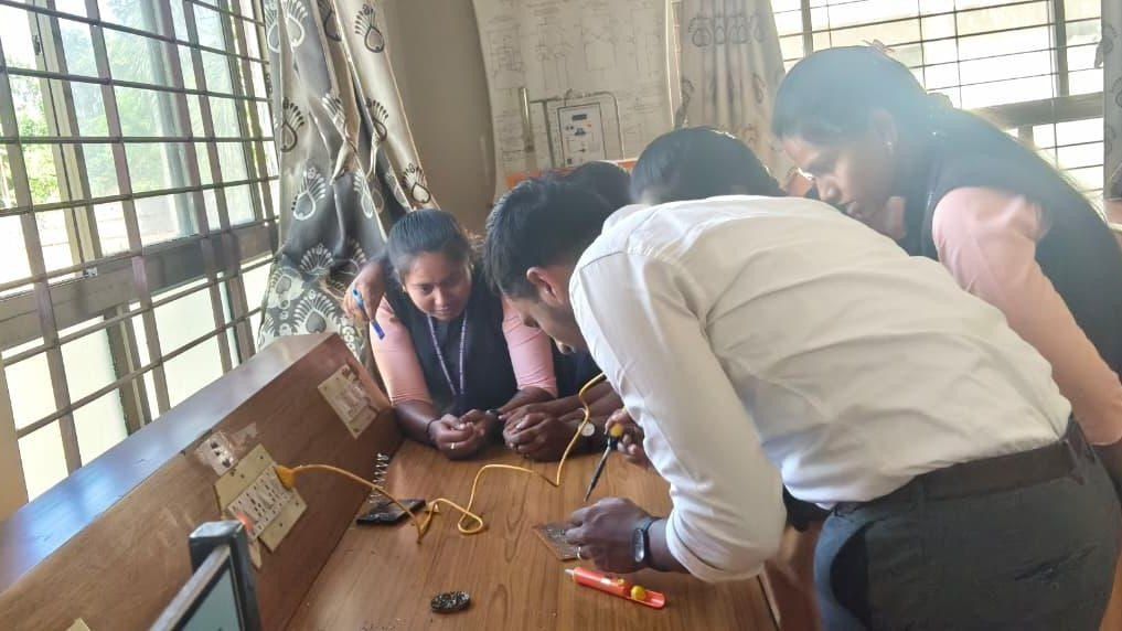 A man in a white shirt and two other people bend over a wooden bench beside a window, working on a wired circuit.