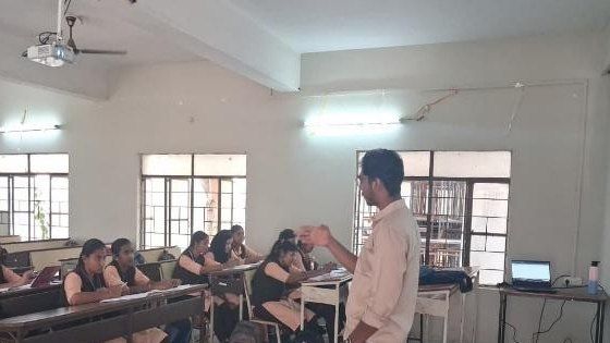 A man in a light shirt gestures while speaking to students seated at long desks in a classroom, with a laptop on a desk at the right.