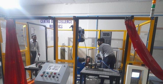 Two people in the robotics cell with its caged robot and a control panel marked HMI, under a Centre of Excellence Robotics sign.