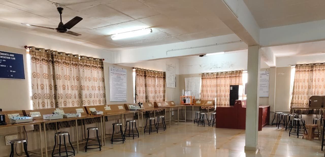 Benches and stools lined up along curtained windows in a hall with a pillar, with a ceiling fan above and a blue name board at the left edge.