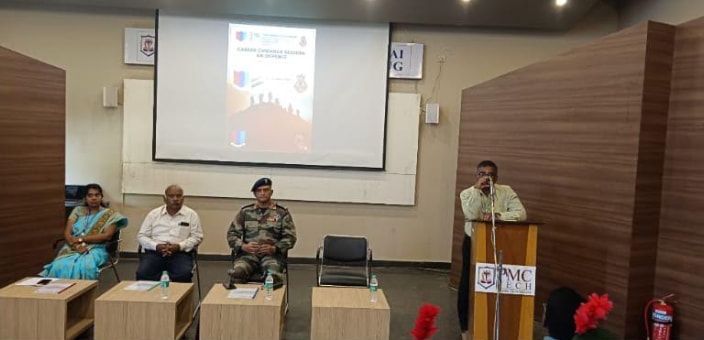Three people seated at small tables at the front of a hall, a poster on the screen behind them and a lectern marked PMC Tech to the right