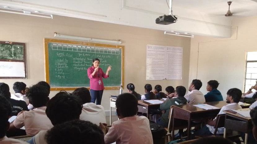 A woman in a magenta kurta stands at a green chalkboard speaking to students seated at desks, with a notice board and a ceiling projector behind her.