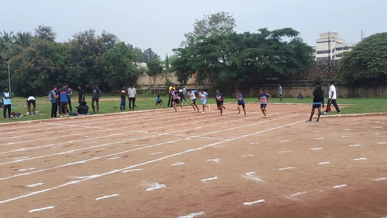 Sprinters just off the blocks on a red earth track marked with white lines, officials and onlookers beside it and trees beyond