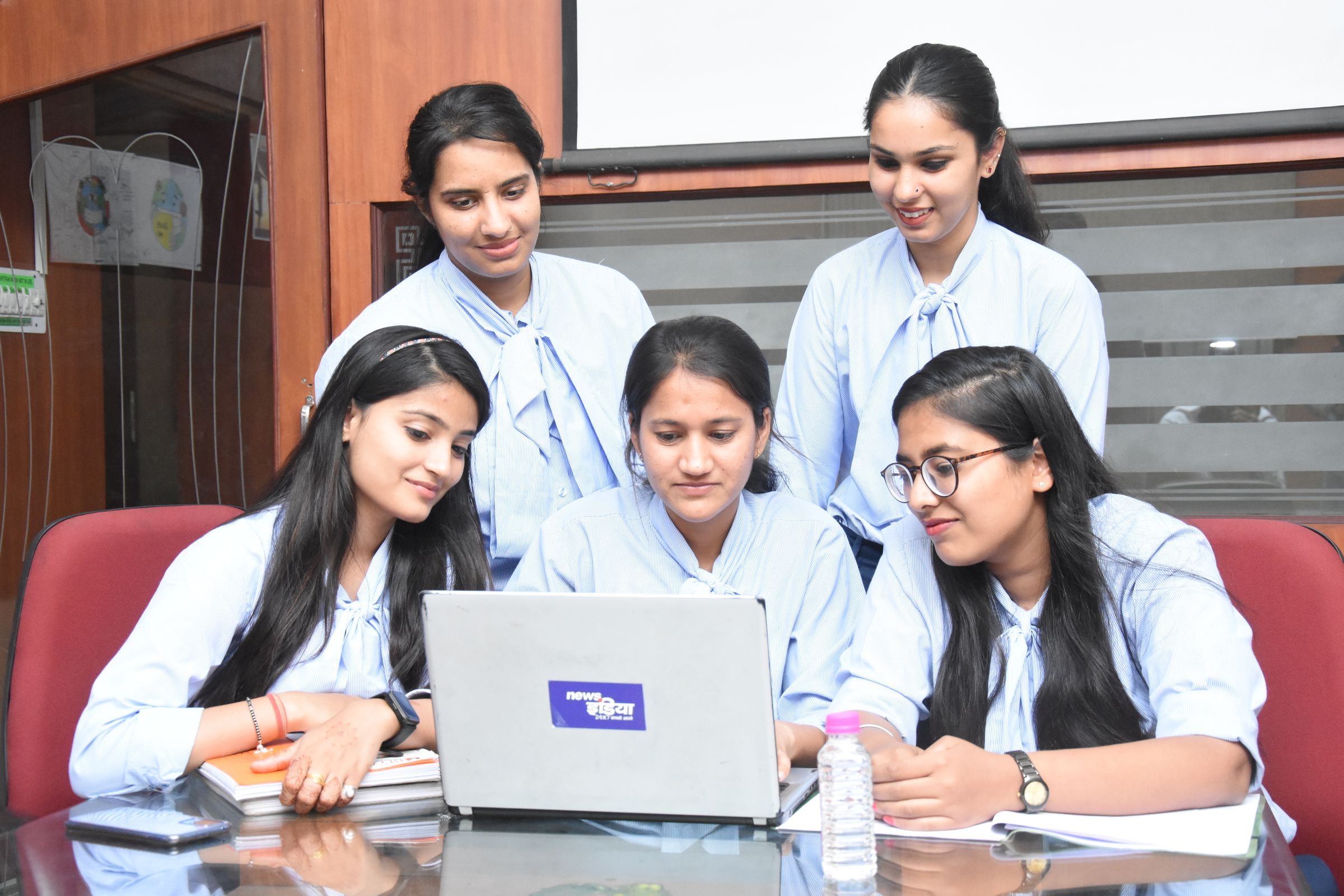 Four students in matching uniforms gathered around a laptop, reading and taking notes