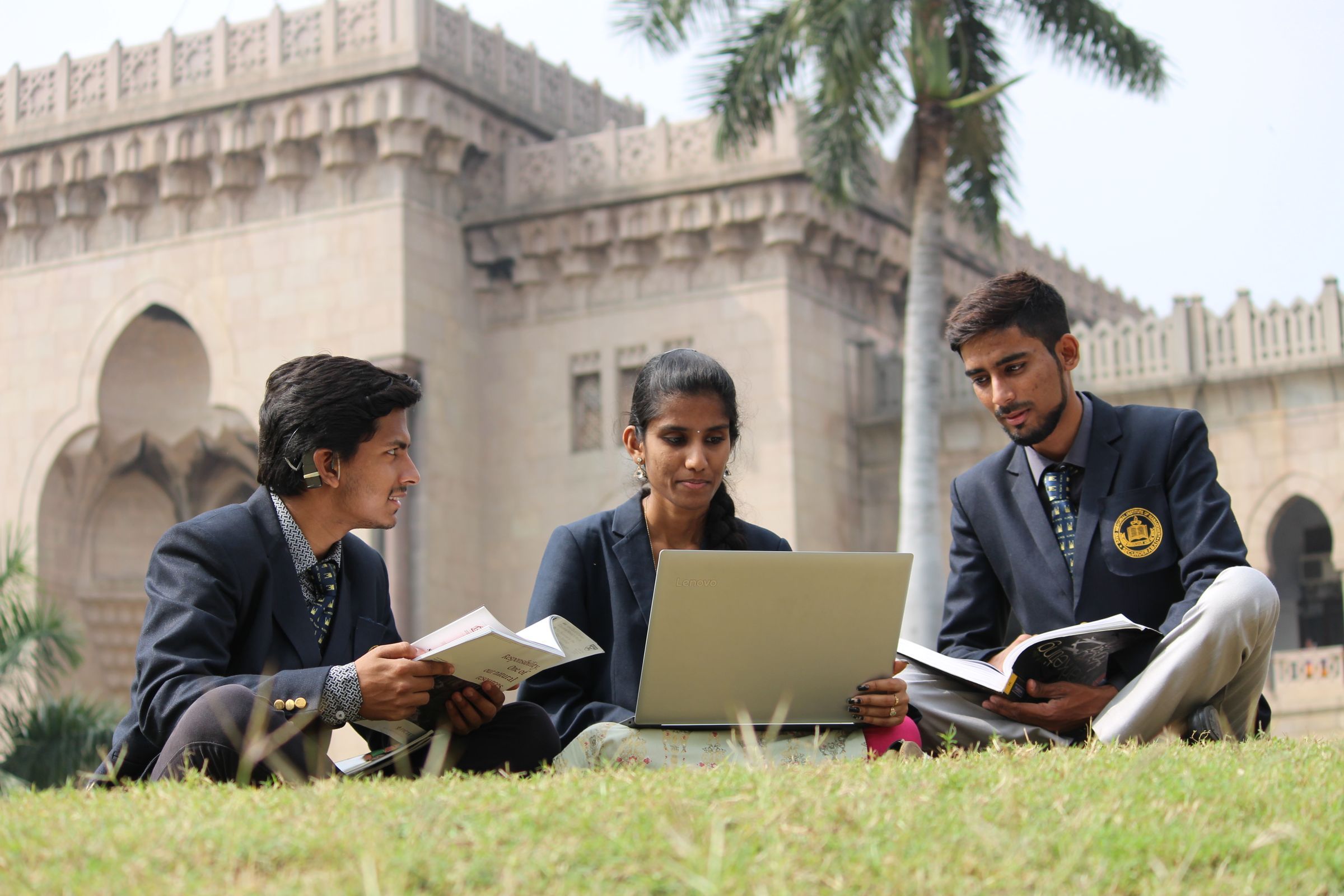 Three students in blazers sitting on a lawn with books and a laptop, in front of a stone building