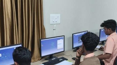 Students in peach-coloured shirts sit at desktop computers beside a curtained window in a computer laboratory.