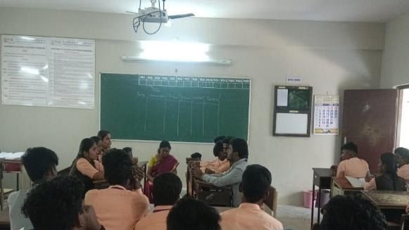 Students sit in rows facing the front of a classroom, with a green board and a notice board on the wall.